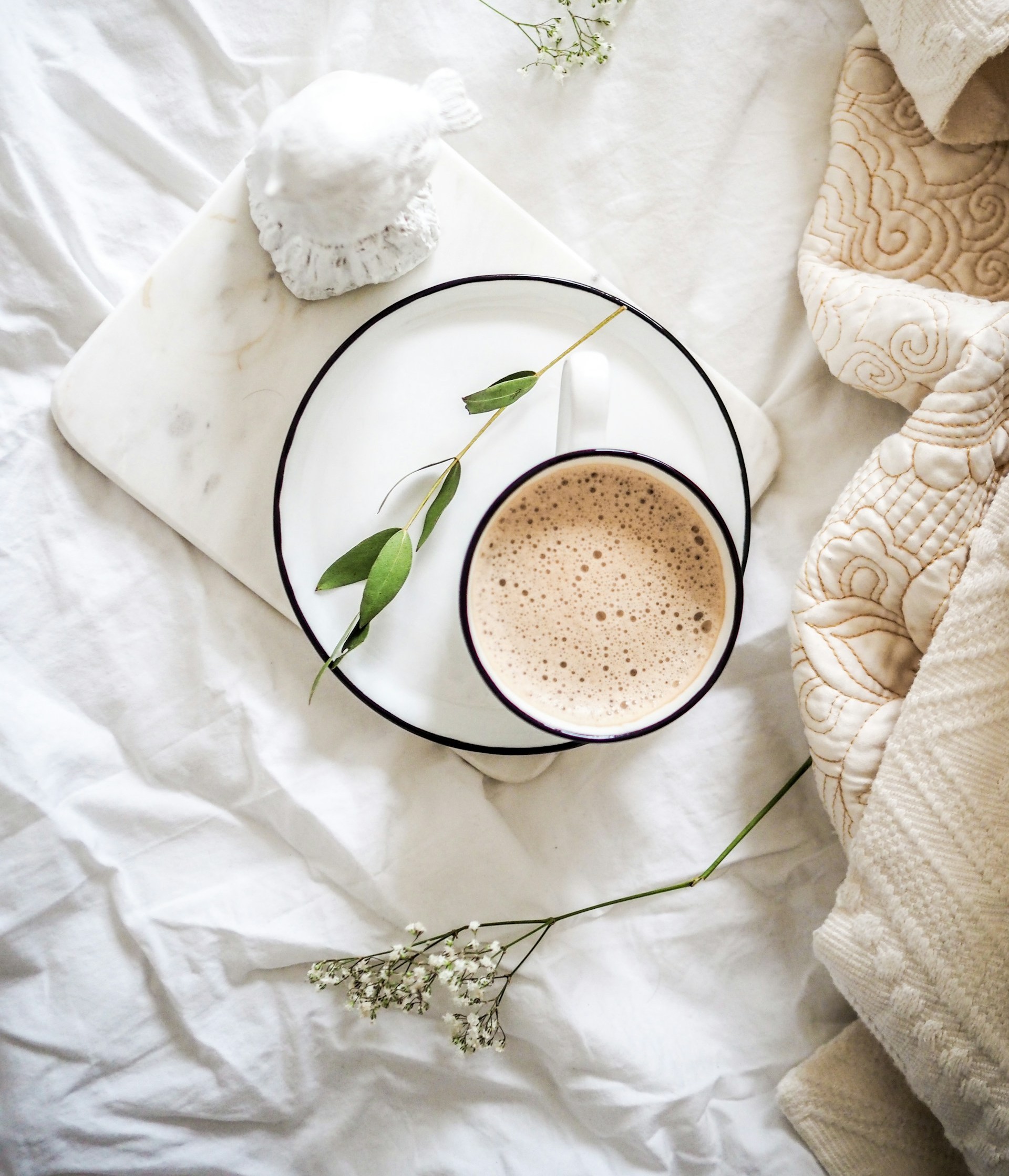Top-down view of a latte in a white cup on a marble tray over white bed linens with a bird figurine and green leaves